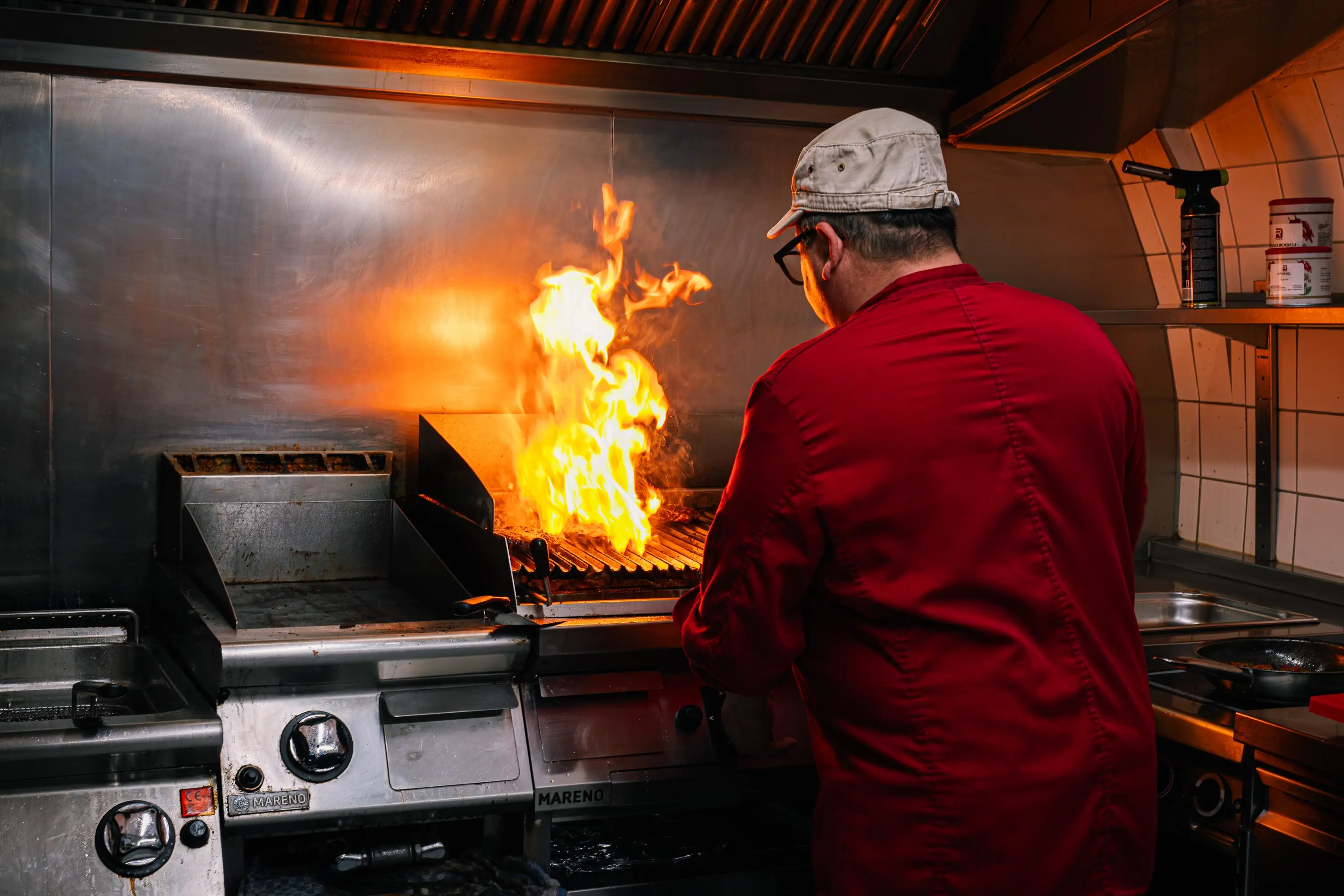 Chef in a red jacket at the grill of a restaurant kitchen, a tall flame shooting up