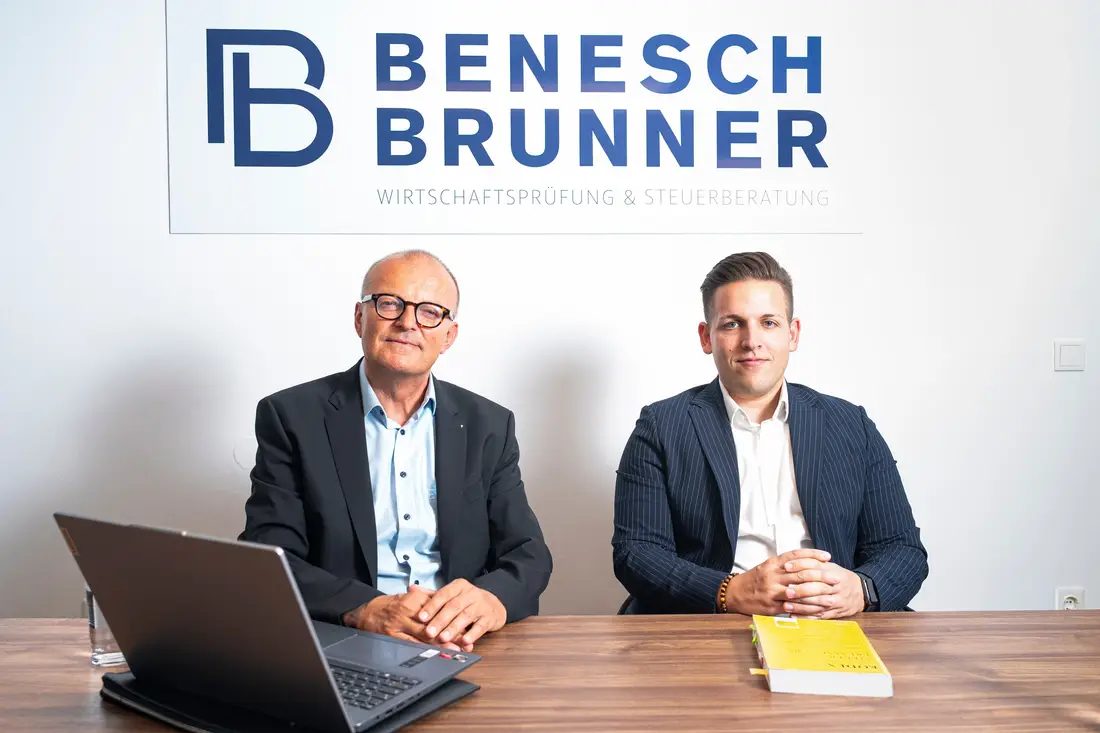Two men in suits at a meeting table in front of the Benesch Brunner logo