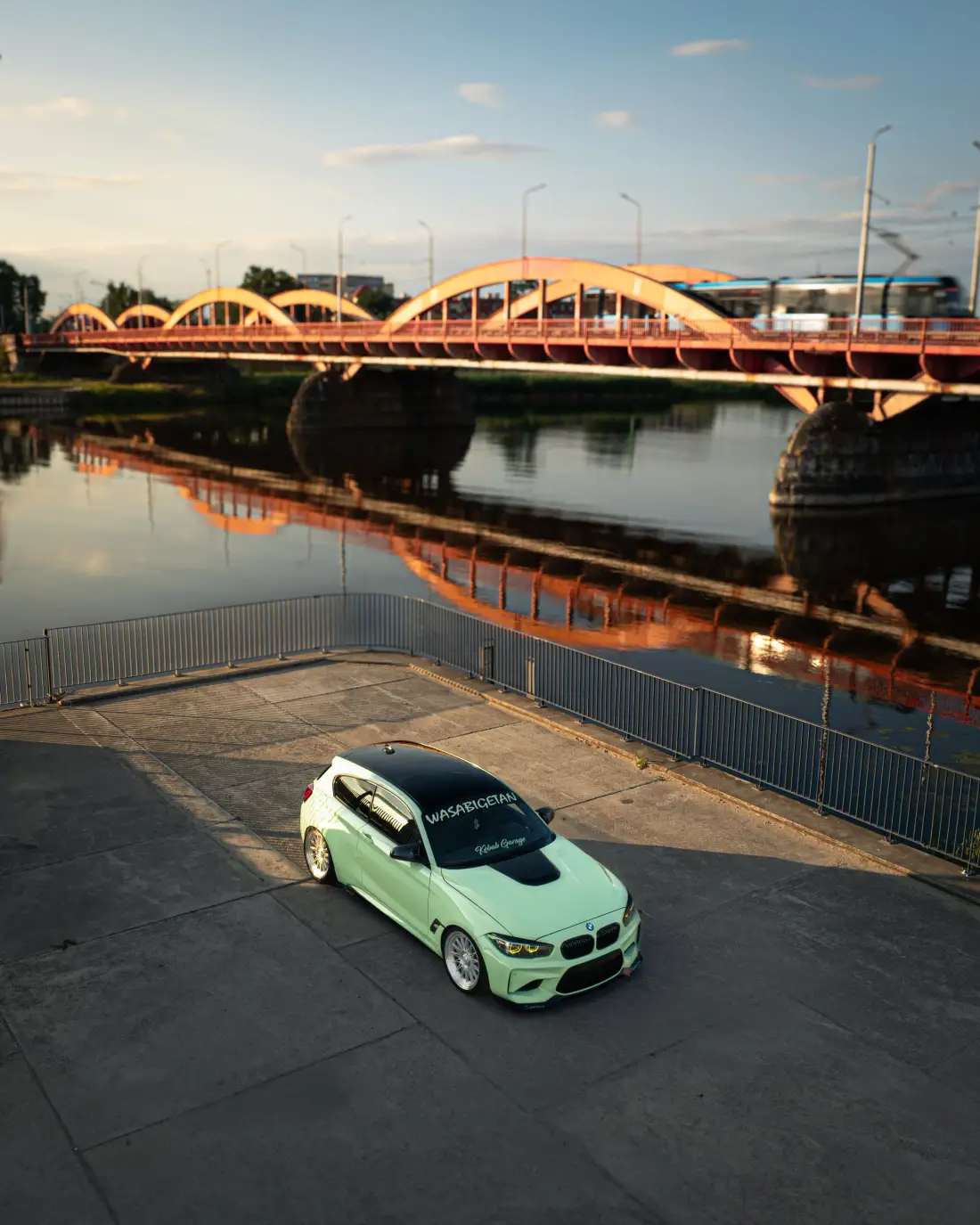 Mintgrüner BMW von oben am Flussufer, dahinter eine rote Bogenbrücke