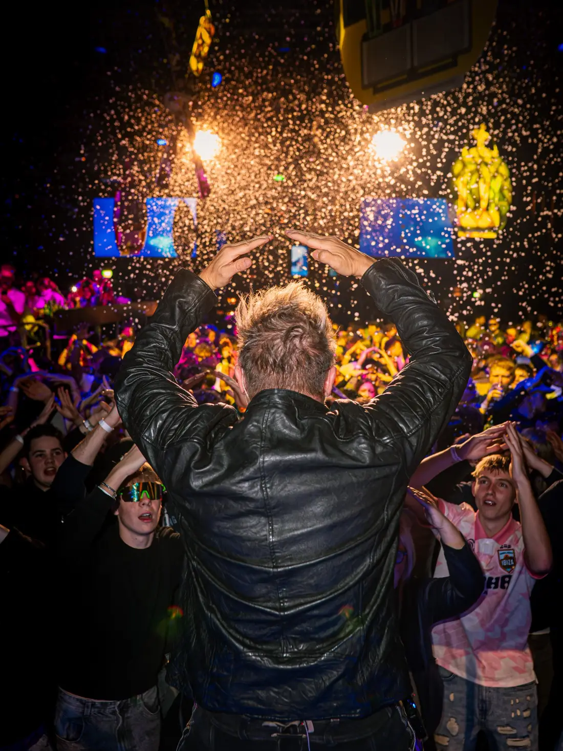 Performer seen from behind forming a heart with his arms, the audience in confetti in front