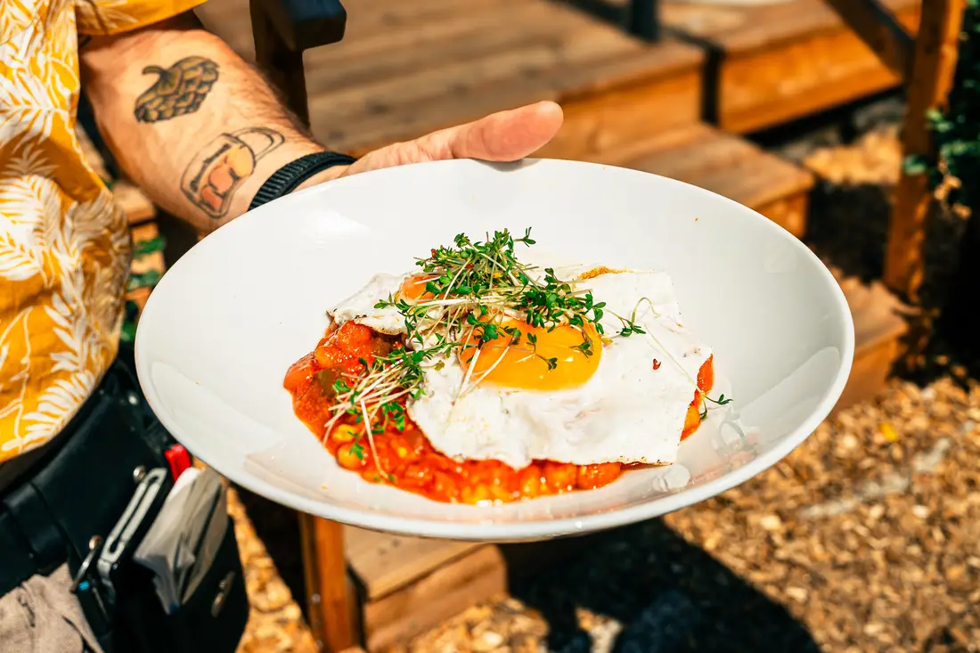 Teller mit Spiegelei auf Tomatensauce und Kresse, gehalten von einem tätowierten Arm