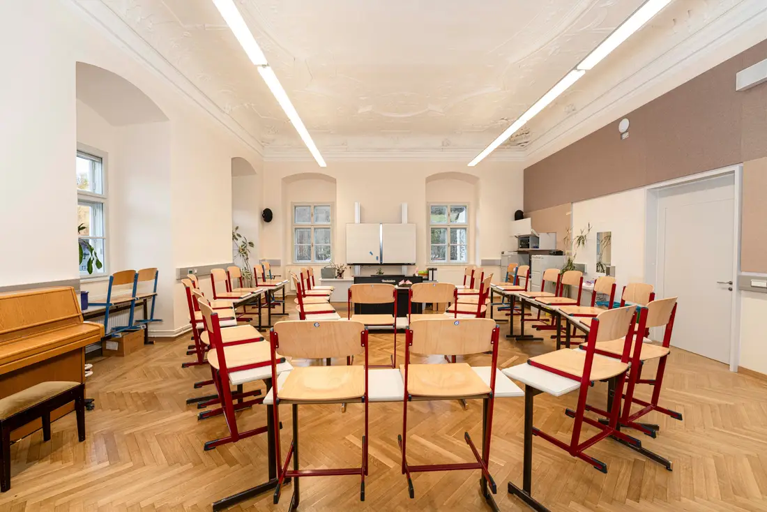 Klassenzimmer mit Stuckdecke, Fischgrätparkett und roten Schulmöbeln