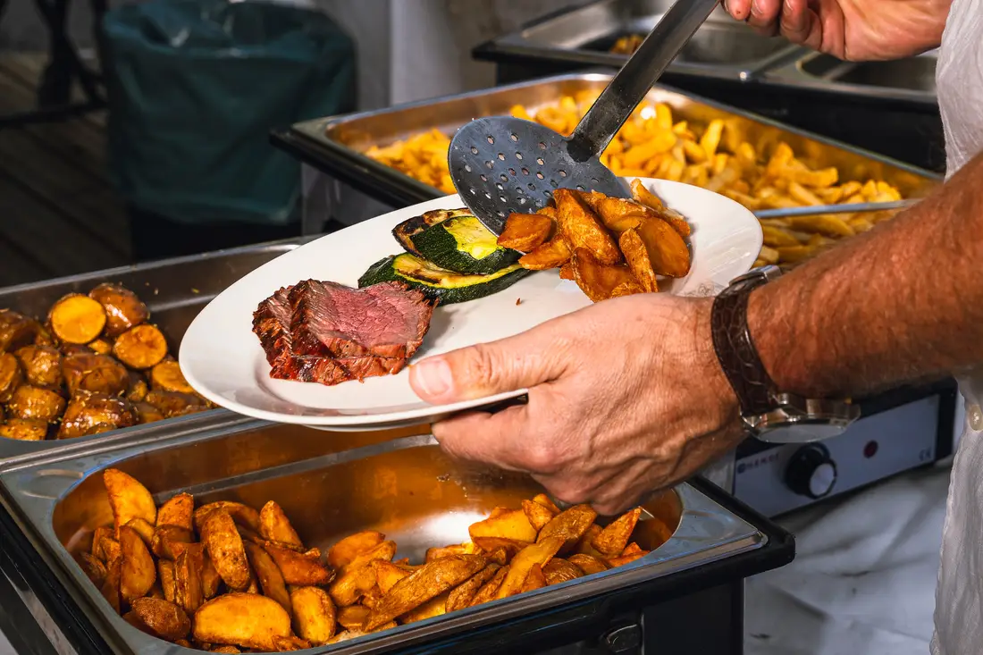Am Buffet wird ein Teller mit Fleisch, Gemüse und Kartoffelspalten angerichtet