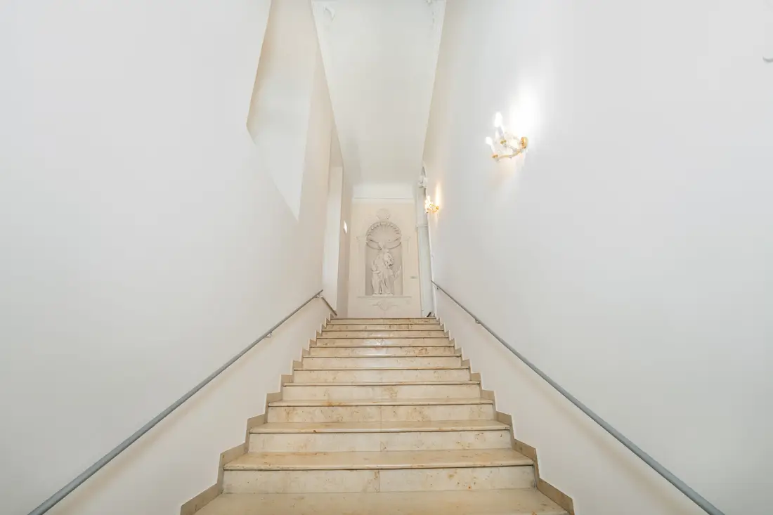 Breite Steintreppe in einem weißen historischen Stiegenhaus mit Wandleuchte