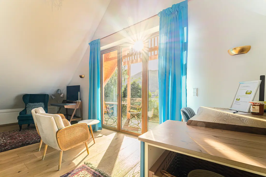 Helles Zimmer mit Holzmöbeln, blauen Vorhängen und Balkontür, durch die die Sonne scheint