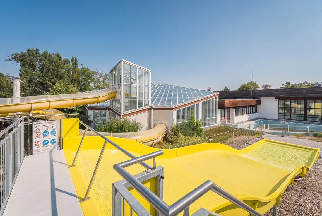 Breite gelbe Wasserrutsche in einem Bad in Krems, dahinter eine Schwimmhalle mit Glasdach