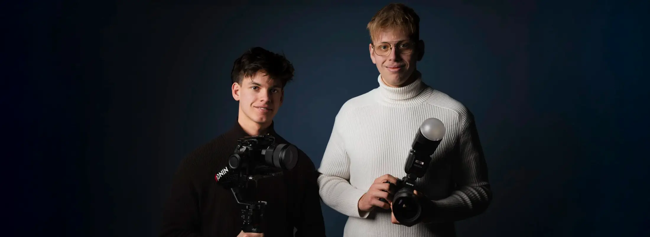 Paul Schneider and Bryan Schmied with cameras against a dark blue background